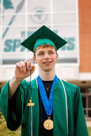 Smiling male graduate in green cap and gown holds tassel while wearing gold medal outdoors.