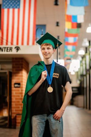 Smiling graduate in green cap holds gown over shoulder wearing gold medal inside school hallway.