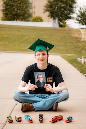 Graduate in green cap sits cross-legged holding childhood photo surrounded by toy trains on pavement.