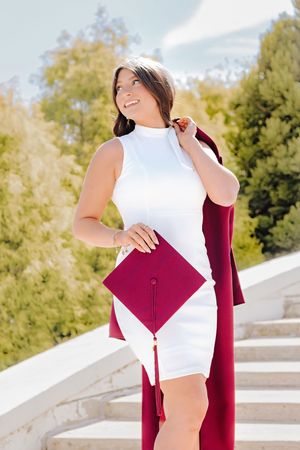 Young woman in white dress holding maroon graduation cap and gown on outdoor staircase.