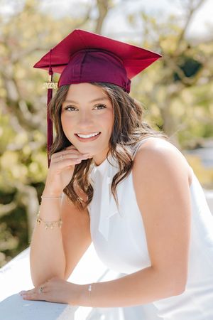 Smiling brunette graduate in white dress and maroon cap posing outdoors in bright sunlight.