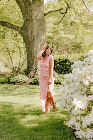 Smiling brunette girl in pink floral maxi dress standing near white blooming shrubs and large tree.