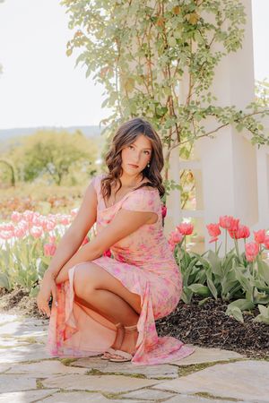Brunette teen in pink floral dress crouching beside tulips near white column in garden.