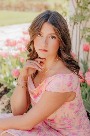 Brunette teen girl in pink floral off-shoulder dress seated among pink tulips in garden.
