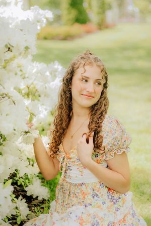 Young woman in floral dress sitting beside large white flowering bush in lush garden. Hershey Gardens Senior Pictures