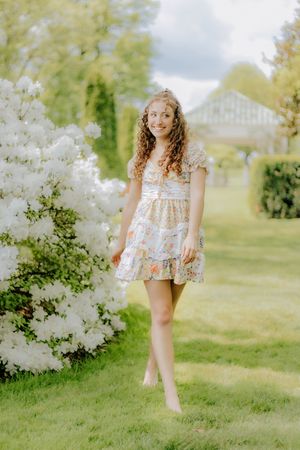 Young woman in colorful floral mini dress walking barefoot near white flowering shrubs. Hershey Gardens Senior Pictures