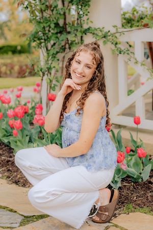 Smiling young woman in blue top and white jeans crouching beside red tulips in garden. Hershey Gardens Senior Pictures