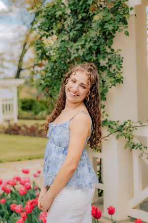 Curly-haired young woman in blue floral camisole smiling near white pergola with red tulips. Hershey Gardens Senior Pictures
