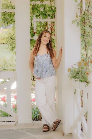 Smiling young woman in blue floral top and white pants leaning on white pergola columns. Hershey Gardens Senior Pictures