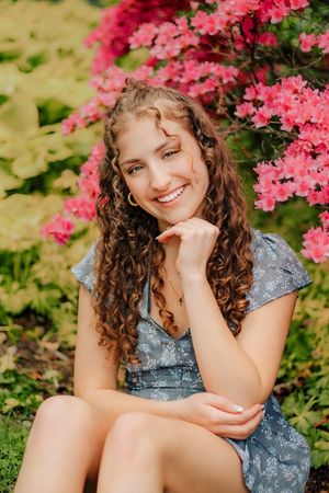 Young woman with curly hair smiling in a blue floral dress among pink azalea blooms. Hershey Gardens Senior Pictures