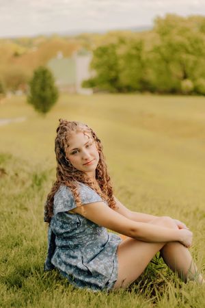Young woman with curly braids in a blue floral dress sitting on a grassy hillside. Hershey Gardens Senior Pictures