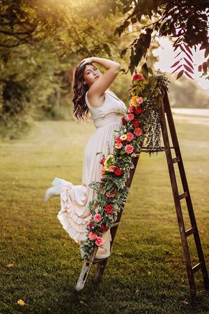 Elegant young woman in white dress posing on a flower-adorned wooden ladder in a sunlit garden. Hanover PA Spring Senior Pictures