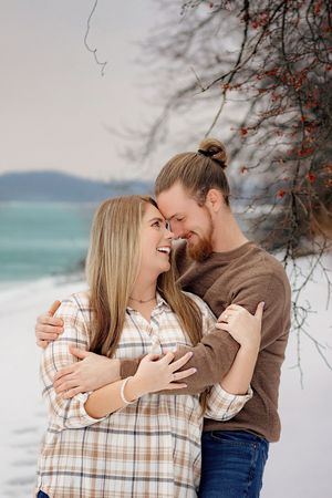 Happy couple embracing and laughing together on snowy beach with turquoise water backdrop. Codorus Couples Winter Pictures