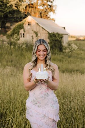 Smiling young woman in pink floral dress holding 16th birthday cake in field near rustic barn.