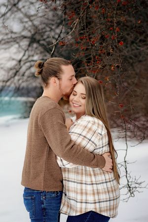 Man kissing smiling woman on cheek while embracing in snowy winter landscape with red berries. Codorus Couples Winter Pictures