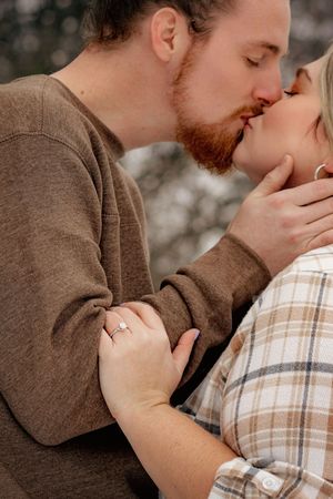 Couple sharing a romantic kiss outdoors, woman displaying an engagement ring while man tenderly holds her face. Codorus Couples Winter Pictu