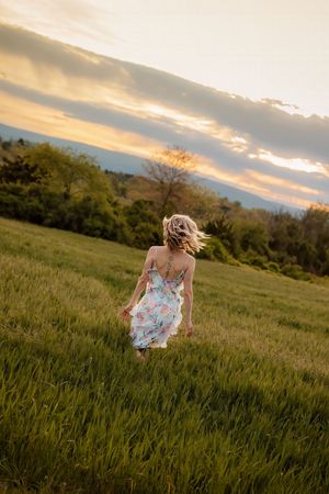 Woman in floral sundress walking away through a lush green field during a dramatic sunset. Gettysburg Senior Pictures