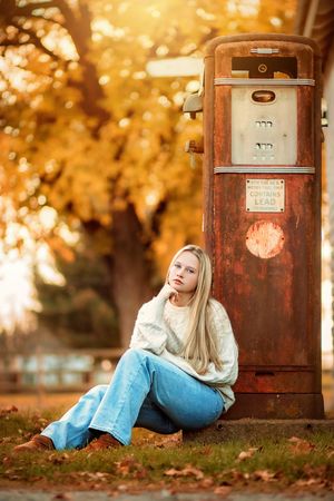 Blonde teenage girl in white sweater and jeans sitting by vintage rusty gas pump in fall.