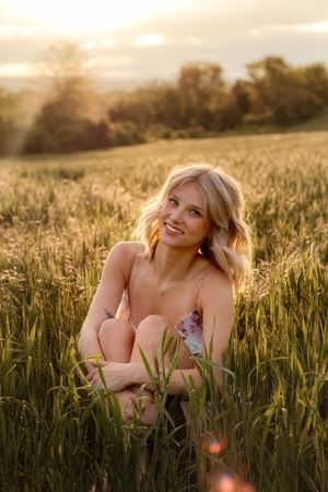Smiling blonde woman sitting in tall green grass during golden hour for outdoor senior photos. Gettysburg Senior Pictures
