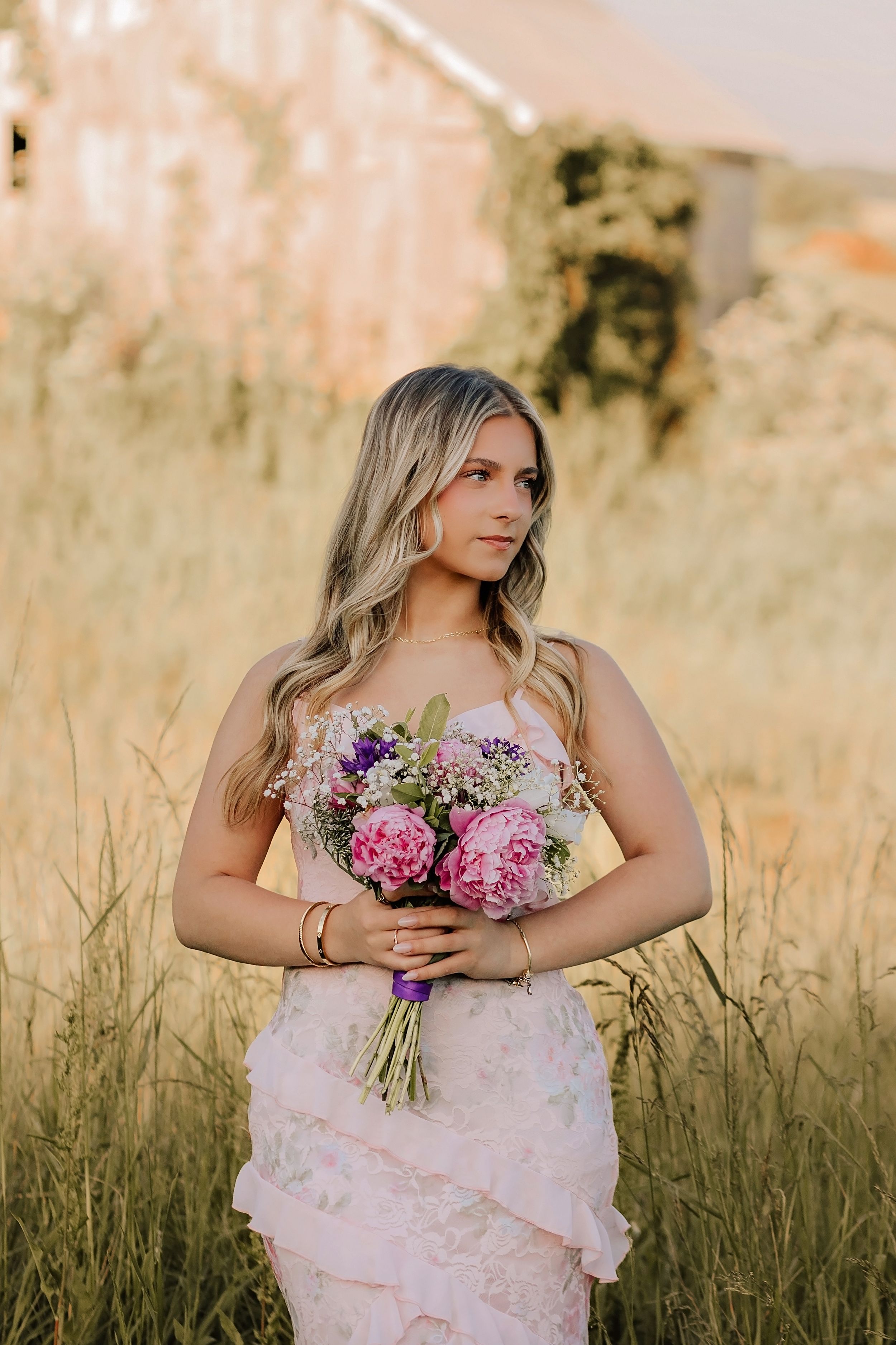 Blonde woman in pink floral gown holding bouquet of pink and purple flowers in golden hour field.