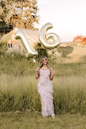 Young woman in pink floral dress holding gold number 16 balloons in sunlit grassy field.