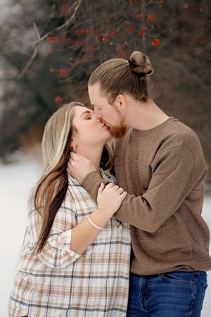 Romantic couple sharing a kiss outdoors in winter snow with red berry branches behind them.Codorus Couples Winter Pictures