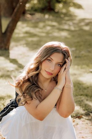 Young woman with wavy brown hair rests hands near face smiling softly in sunny park.