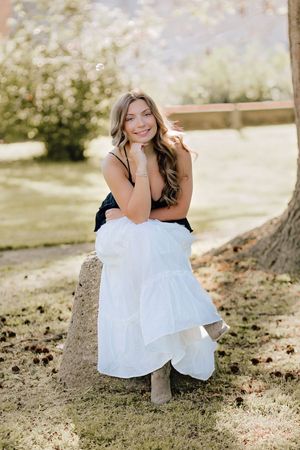 Young woman in black top and white skirt sitting on a rock in a park, smiling for senior portraits. Hanover Pa Spring Pictures