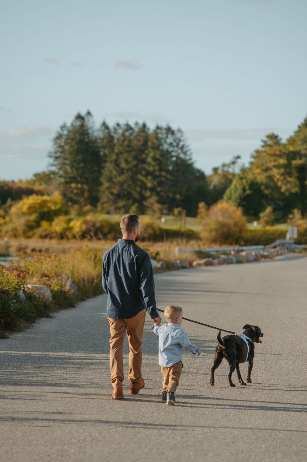 A person and child walk their black dog along a scenic path surrounded by autumn trees and brush at sunset.