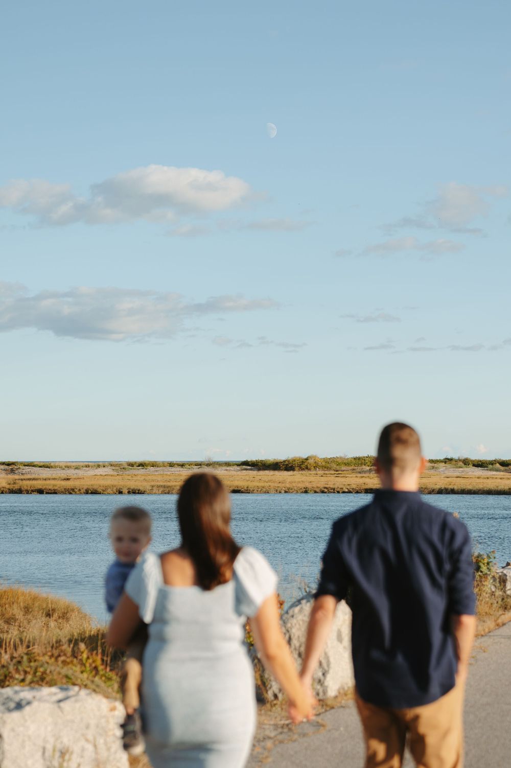 A group of people walking along a coastal path with water and sand dunes visible in the background.