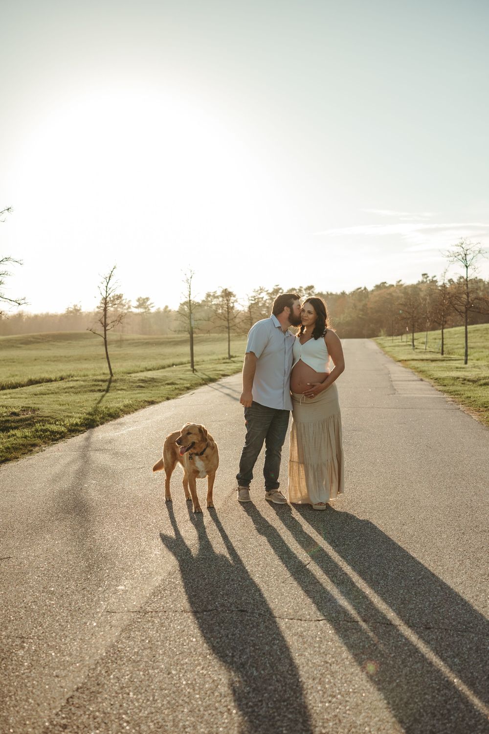 Pregnant couple sharing a kiss on a sunlit road with their dog, surrounded by green fields and trees at golden hour.