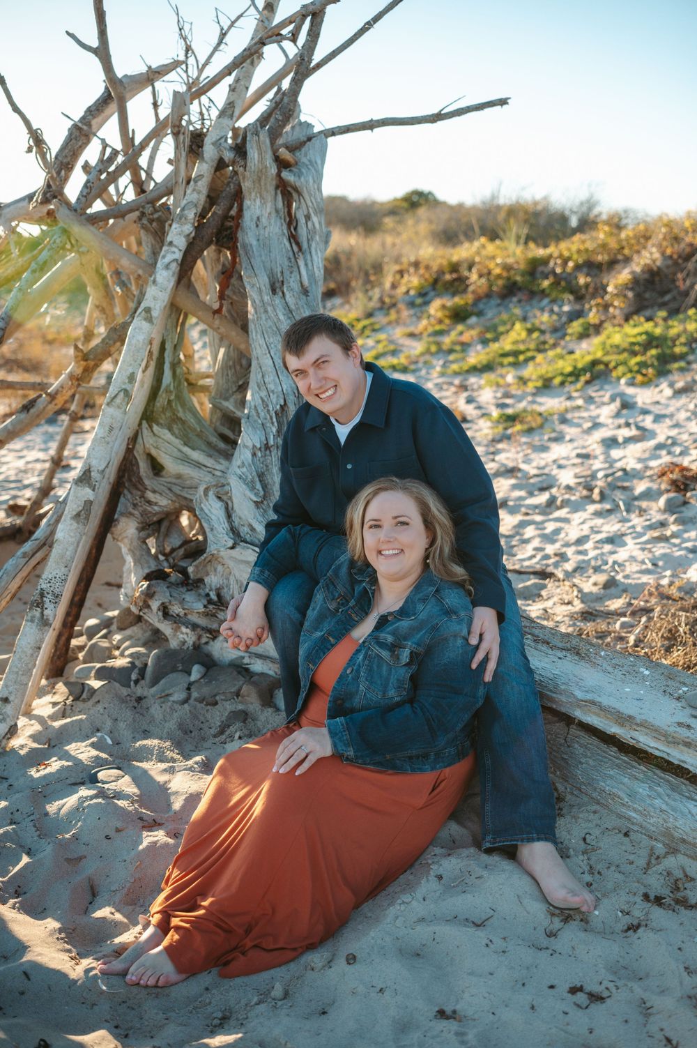 Couple enjoying a romantic moment on a sandy beach with driftwood during golden hour.
