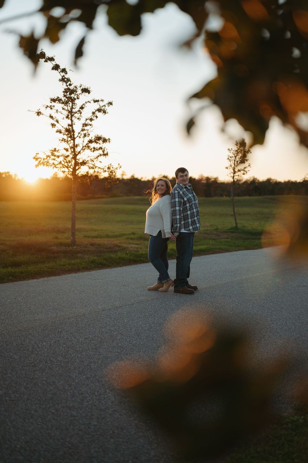 A couple stands together on a country road at sunset while golden light streams through autumn trees in the background.