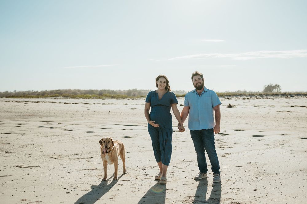 Pregnant couple holding hands walking on beach with dog on sunny day.
