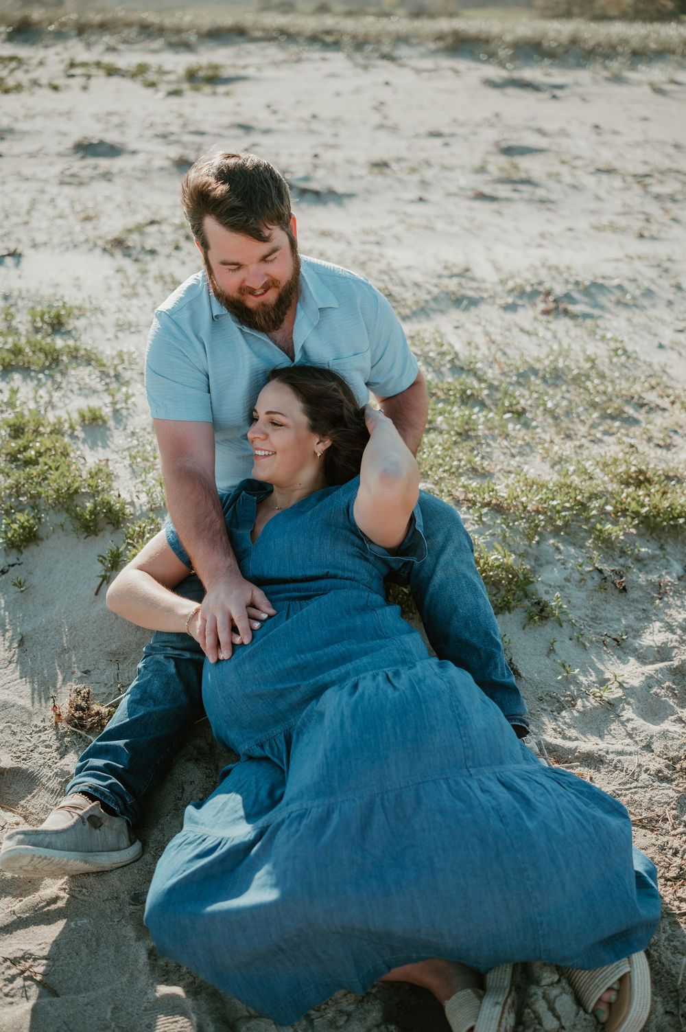 A couple relaxes on sandy beach, woman in blue dress leaning back laughing while bearded man smiles down at her.