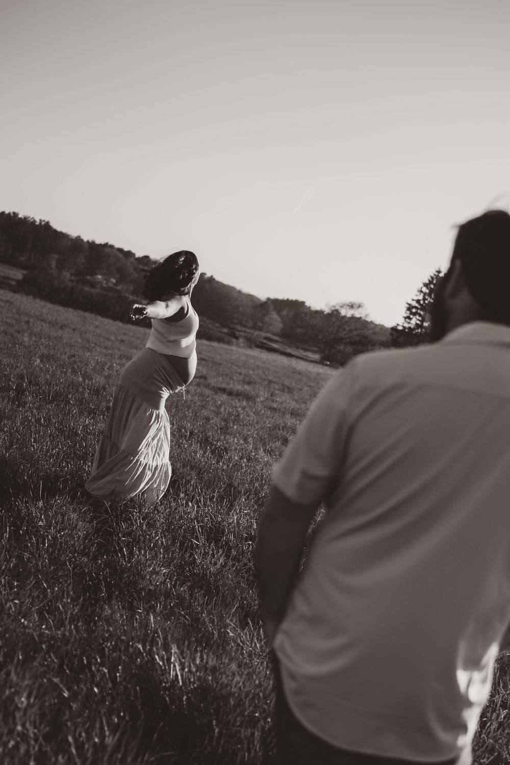Black and white photo of a couple walking through a grassy field at sunset, woman in flowing dress turning back.