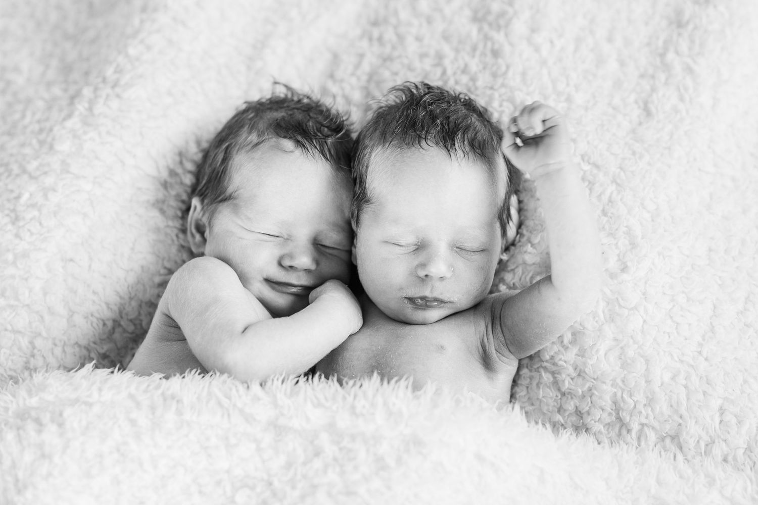 Two sleeping newborn babies lying close together on soft white fur blanket in black and white portrait.