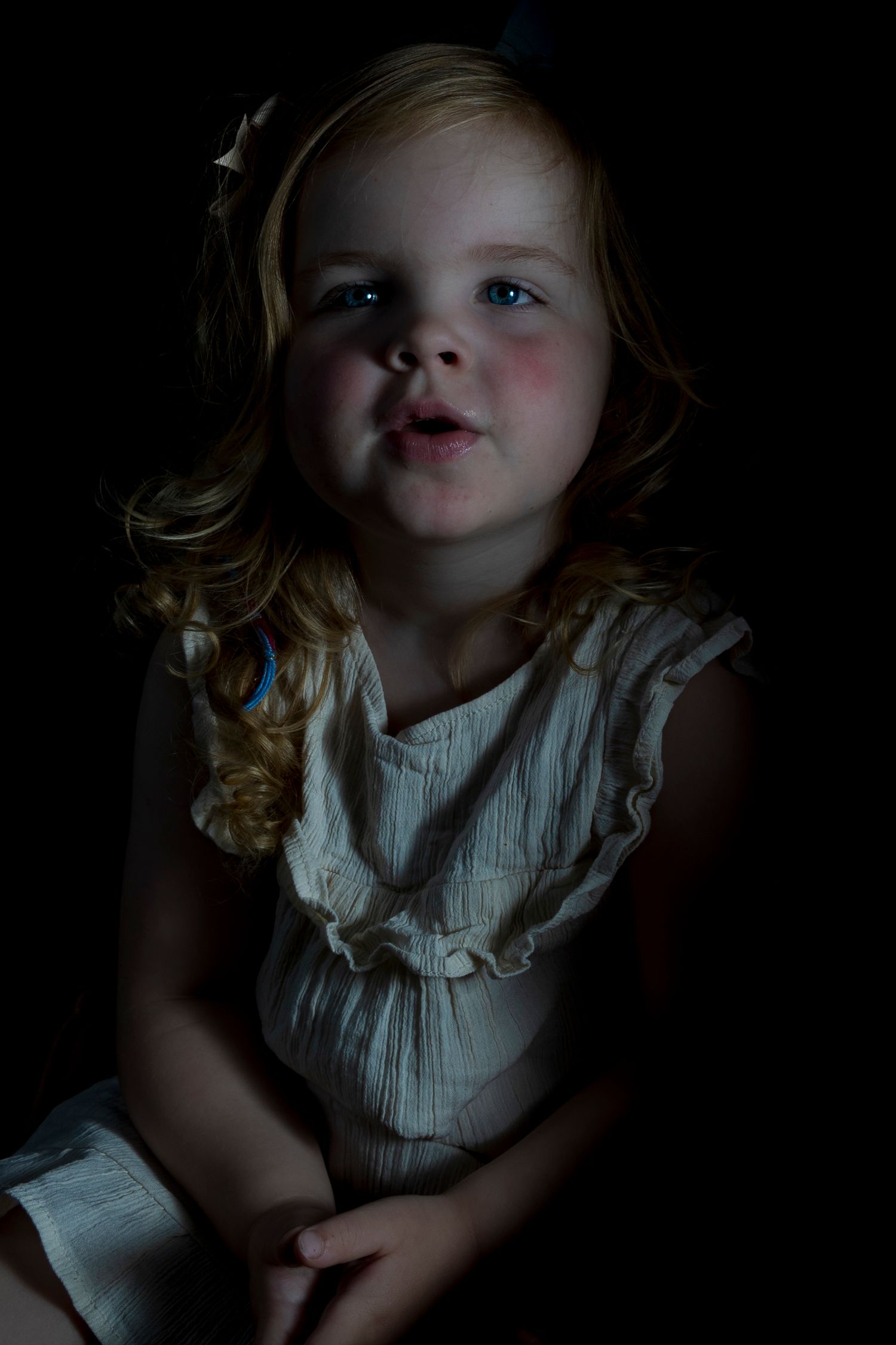 A dramatic low-light portrait of a child in a white dress with blonde hair against a dark background.