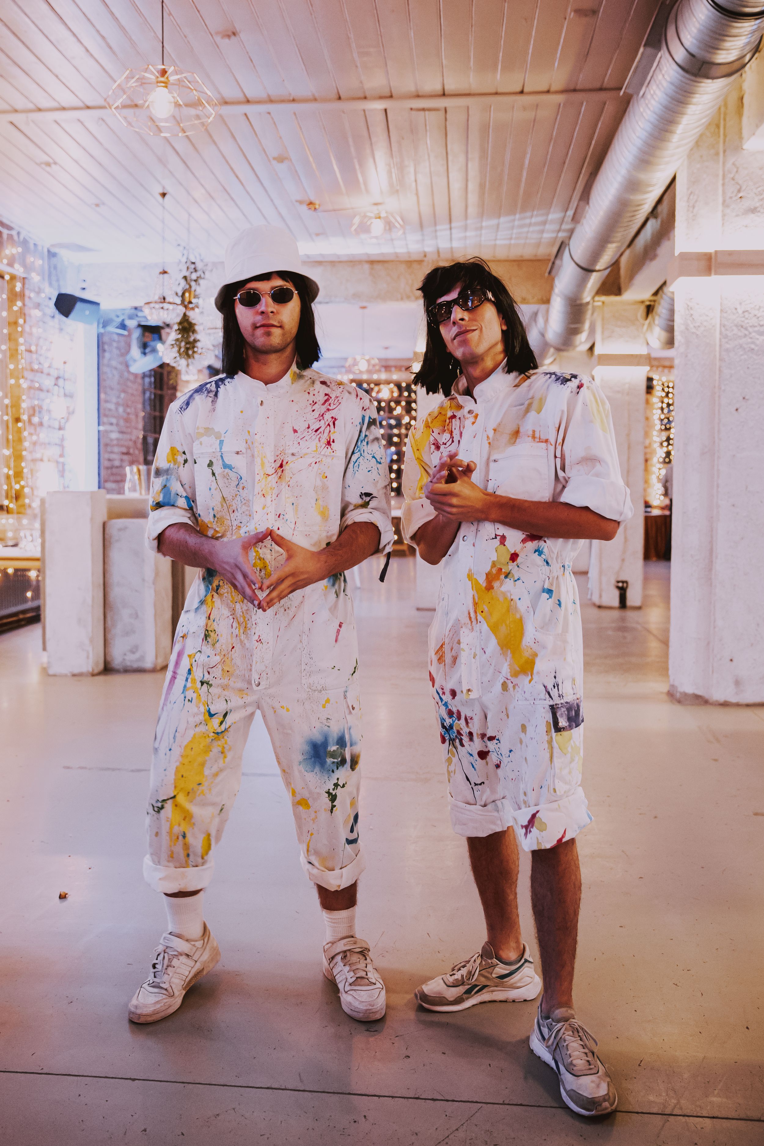 Two people wearing matching paint-splattered outfits pose in an industrial space with string lights.