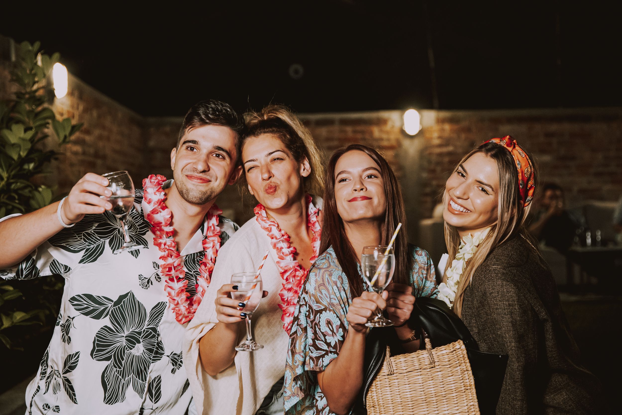 Friends pose with drinks and party props at a nighttime outdoor celebration.