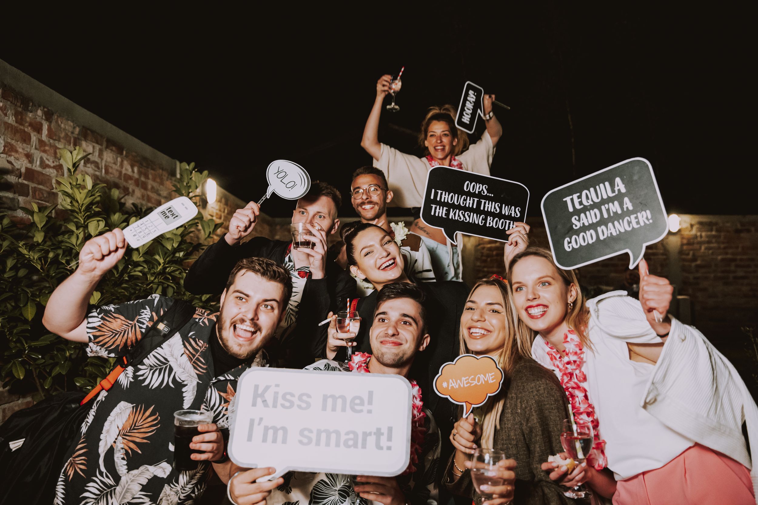 Party guests hold up fun photo booth props and drinks while celebrating at an outdoor nighttime gathering.