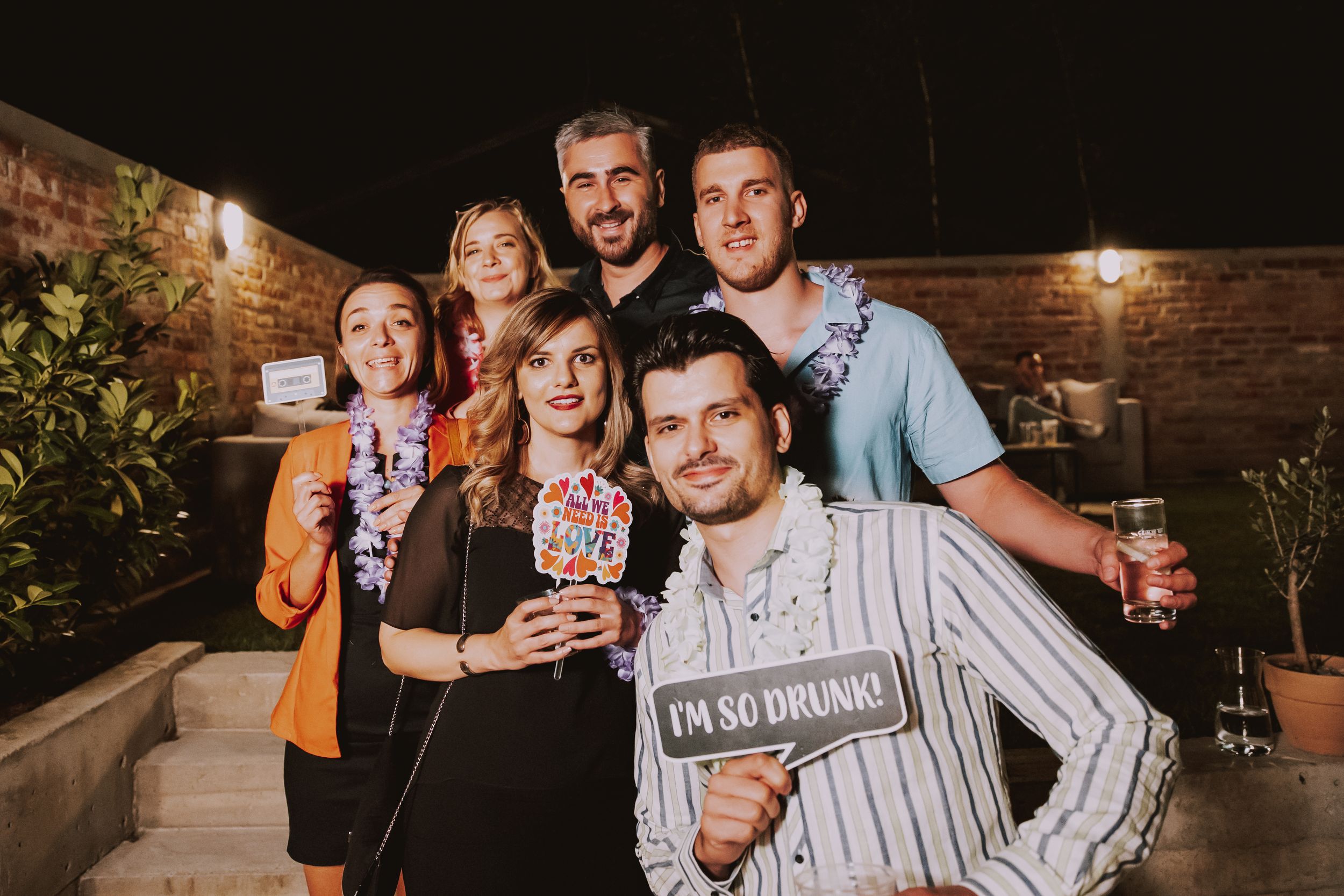 Group celebrating with drinks and photo props against a brick wall backdrop with string lights.