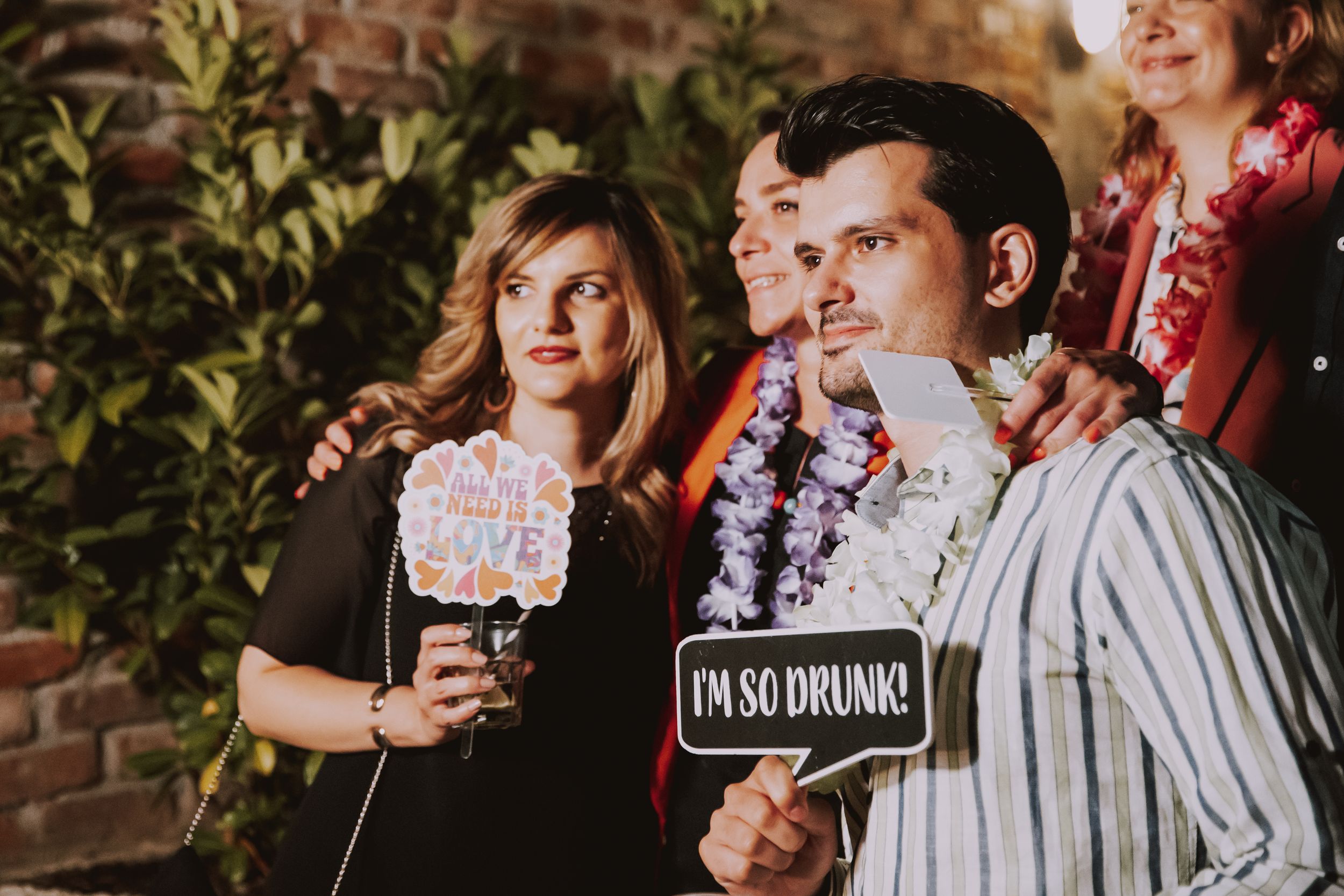 Partygoers hold up humorous signs and wear party leis at an outdoor evening event.