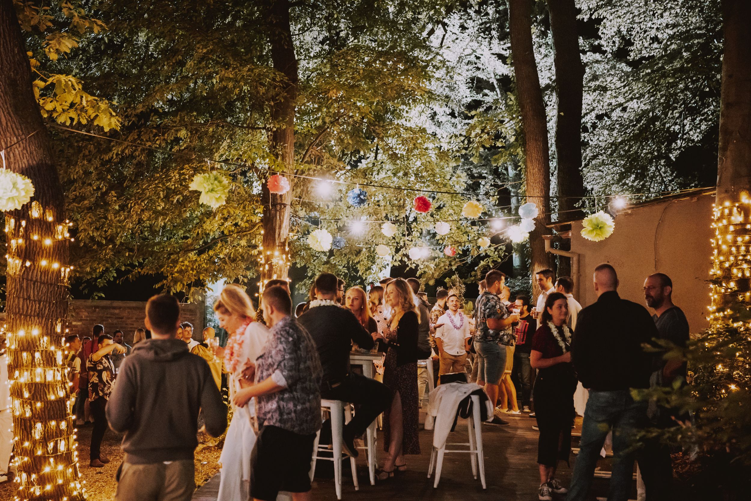 String lights illuminate an outdoor evening party with guests mingling in a garden setting.