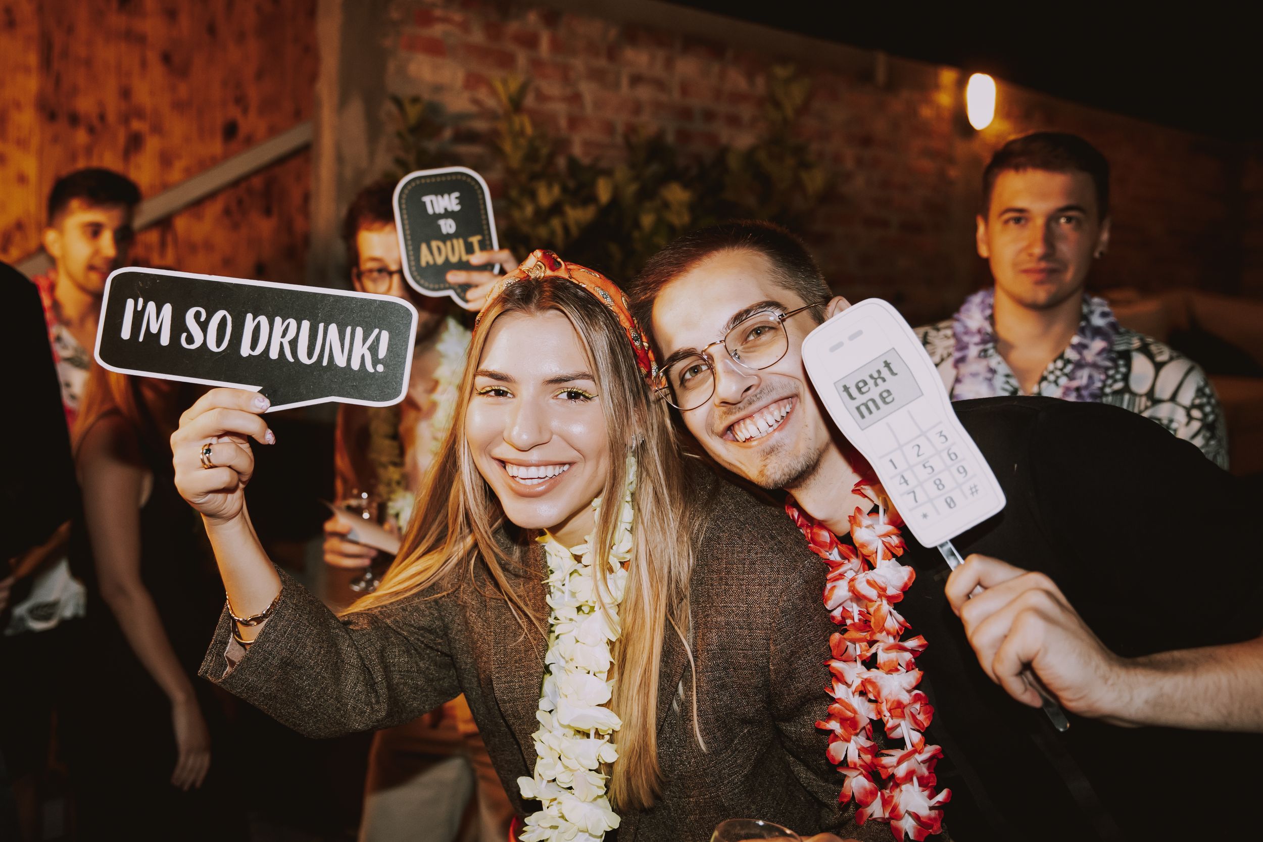 Party guests pose with silly signs and leis while celebrating outdoors at night.