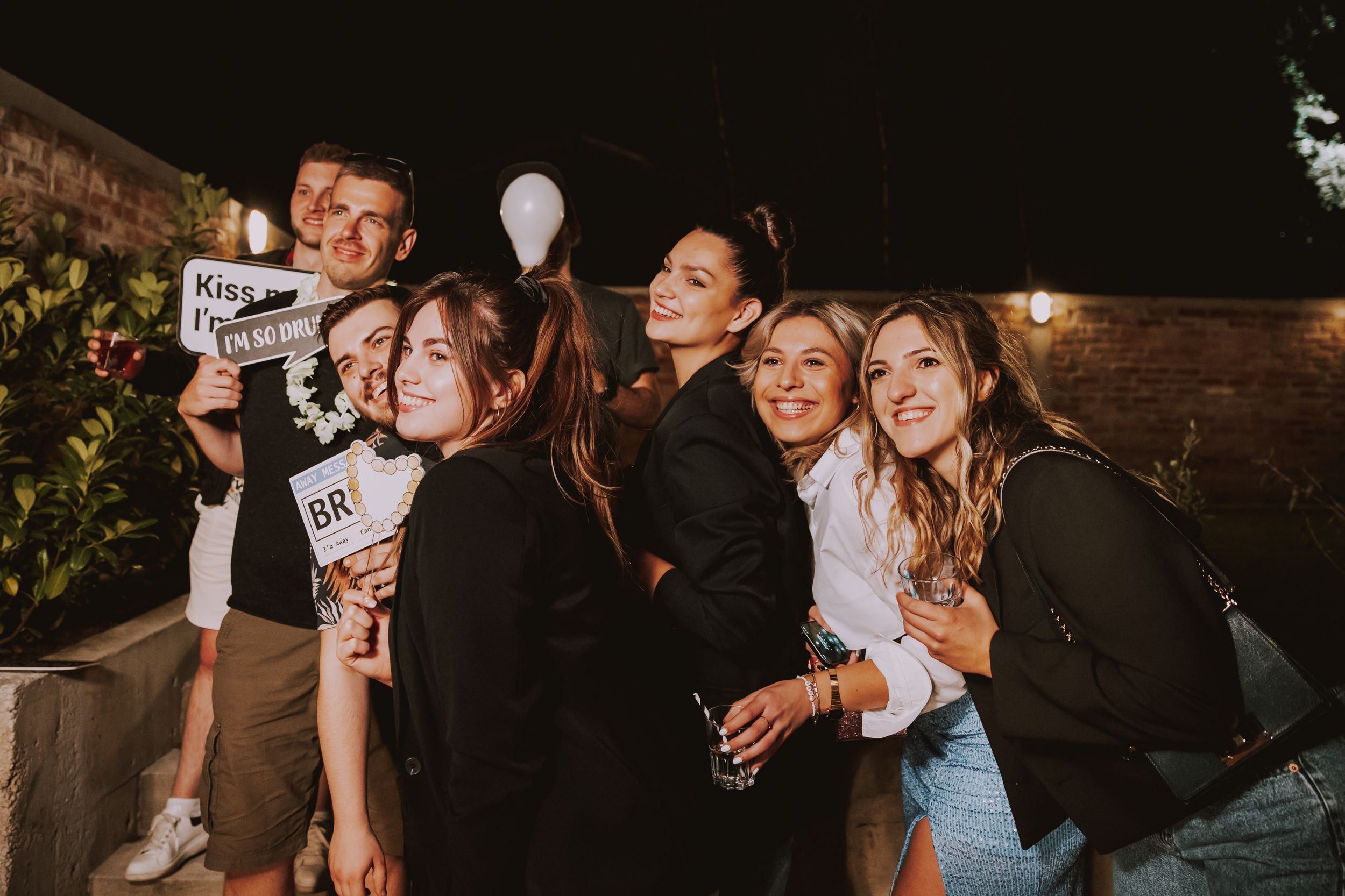 Group poses with photo props and drinks at an outdoor evening celebration.