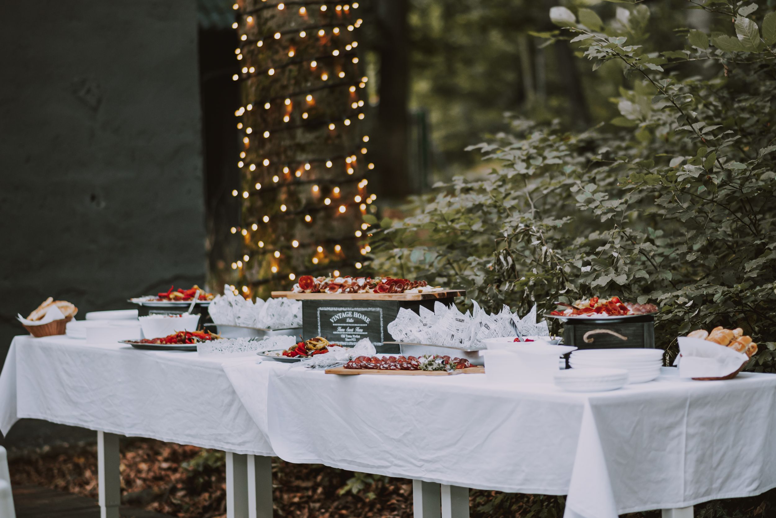 Elegant white tablecloth buffet table decorated with string lights and food displays at an outdoor event.