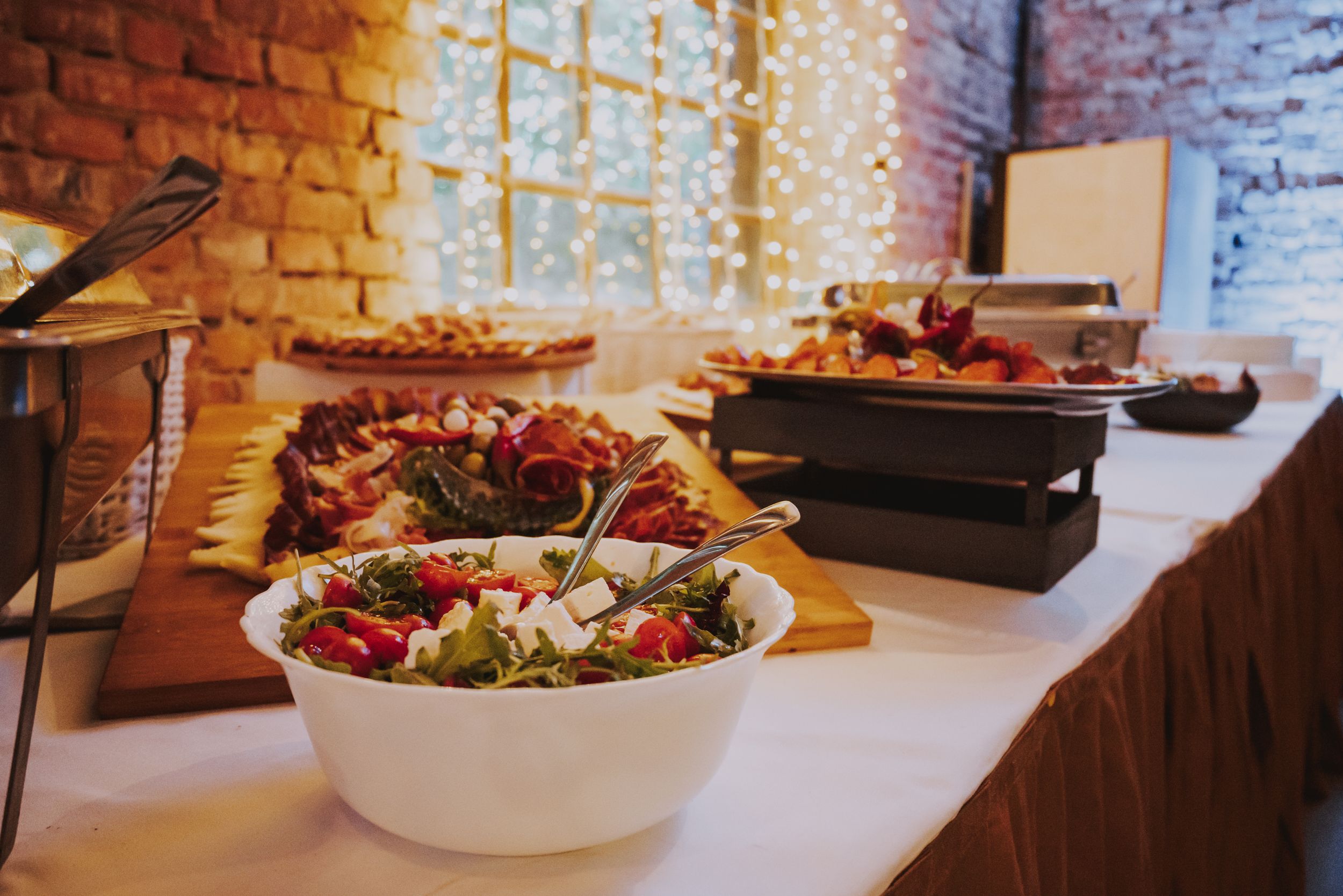 Buffet table with salads and dishes displayed against brick wall with string light backdrop.