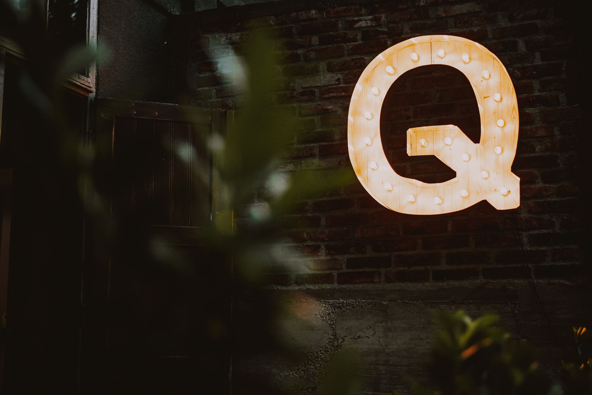 Illuminated letter Q sign mounted on rustic brick wall with plant in foreground.