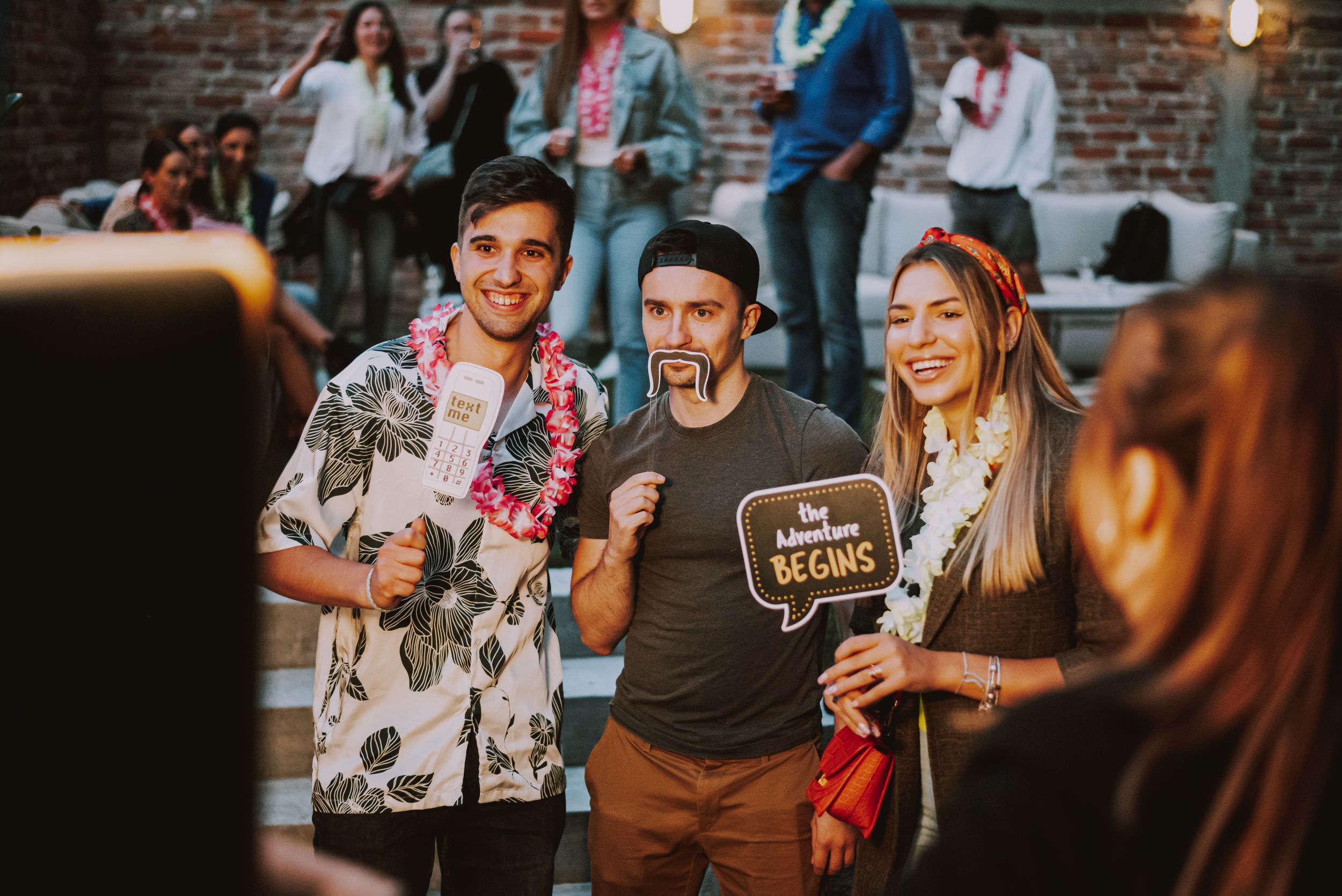 Friends gather for photos at a casual outdoor party with string lights and brick wall backdrop.